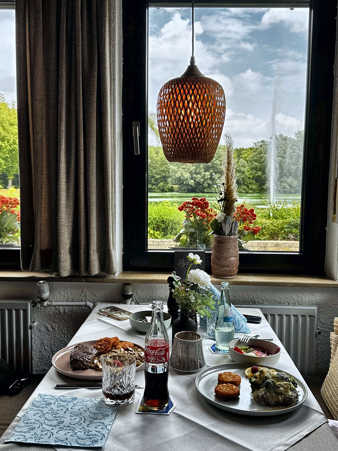 Gedeckter Tisch am Fenster, dahinter Wiese und Baumreihe